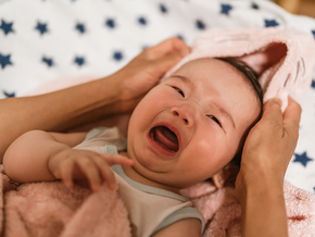 Photograph: crying baby wrapped in pink towel, adult hands gently cradling baby's head