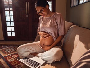 Zwangere vrouw op de bank die haar buik vasthoudt en op een laptop kijkt