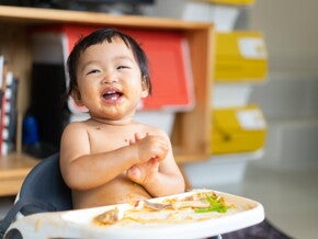 Lachende baby in een kinderstoel met eten op gezicht en tafel