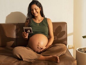 Une femme enceinte assise sur le canapé sourit en regardant une image d’échographie de son bébé