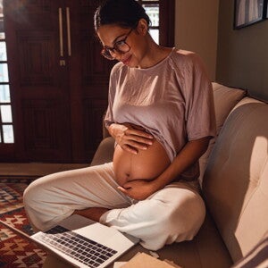 Femme enceinte assise sur le canapé, tient son ventre et regarde un ordinateur portable