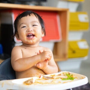Un bébé souriant dans une chaise haute, avec de la nourriture sur son visage et son plateau