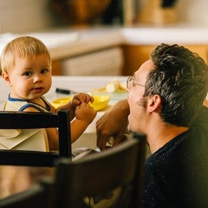 Photographie d'un père agenouillé offrant un jouet jaune à son bébé en chaise haute