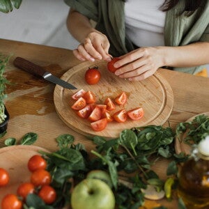 Une femme coupe des tomates sur une planche en bois, entourée de légumes et d’herbes fraîches