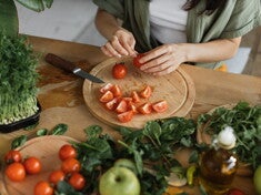 Une femme coupe des tomates sur une planche en bois, entourée de légumes et d’herbes fraîches