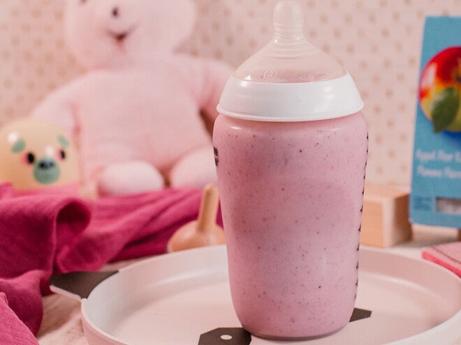 Flesje met roze Aardbeienshake op een dienblad, in een babykamerdecor met speelgoed op de achtergrond.