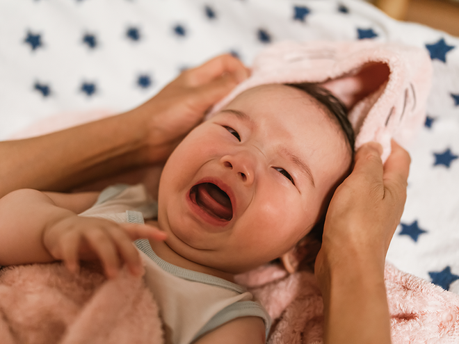 Photograph of crying infant wrapped in pink towel, adult hands gently drying face