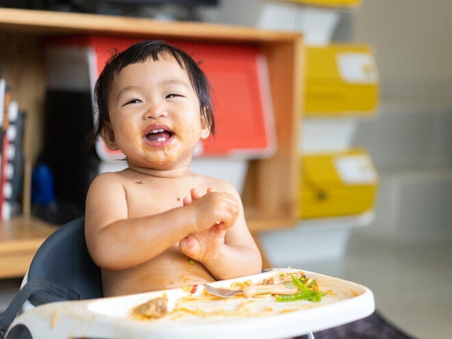 Lachende baby in een kinderstoel met eten op gezicht en tafel