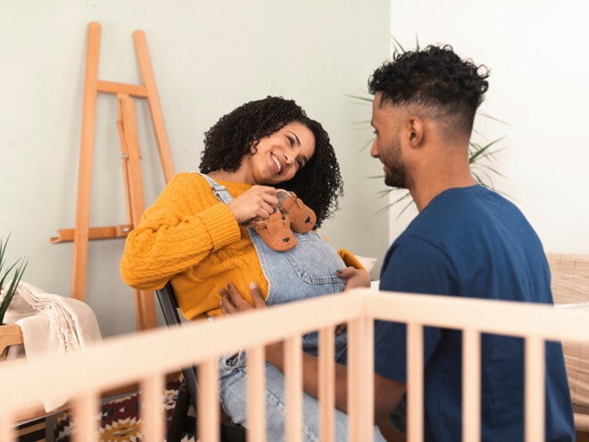 Zwangere vrouw die lachend baby­schoentjes op haar buik houdt terwijl haar partner voor haar knielt